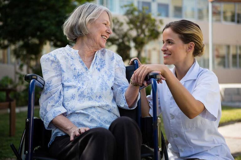 Two older adults receiving help from a caregiver while reviewing a pill organizer in a comfortable residential living room.